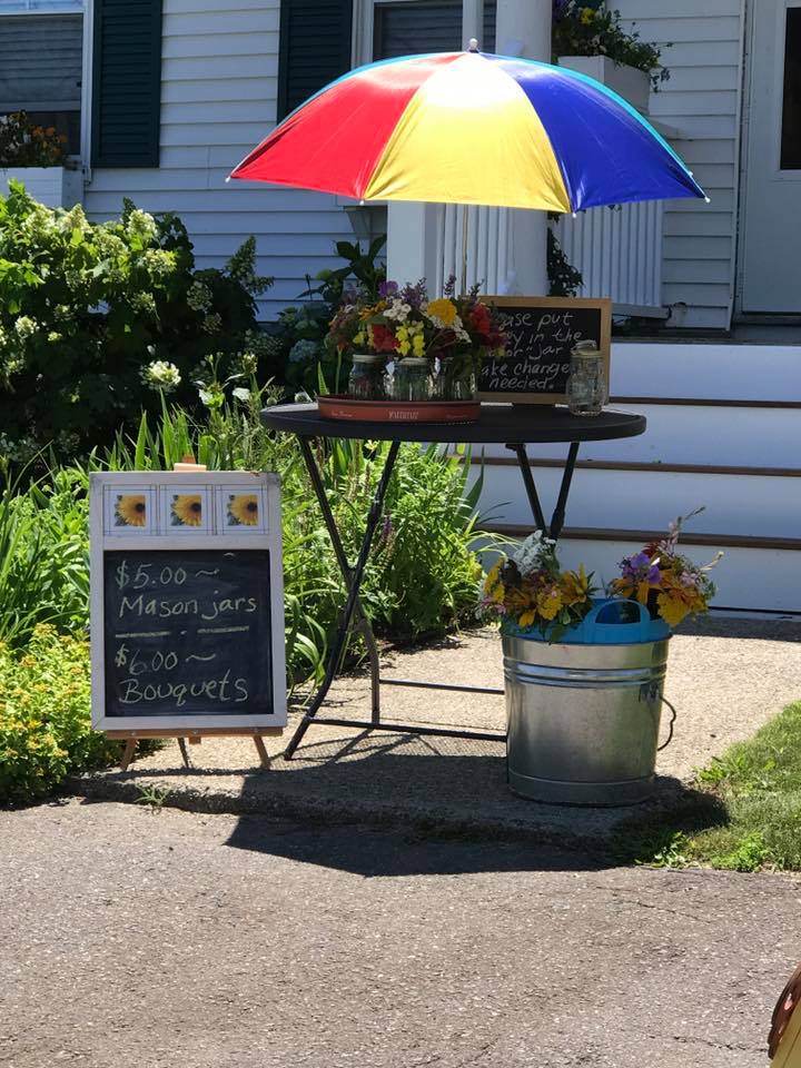 Flower stand with a red, yellow, and blue umbrella and a sign displaying prices for mason jars and bouquets