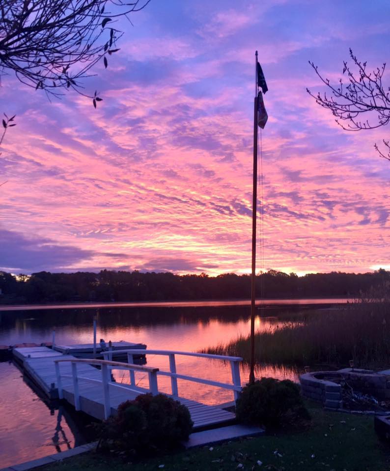 View of the lake, dock, and flag overlooking a multicolored sky in blue, purple, pink, and orange